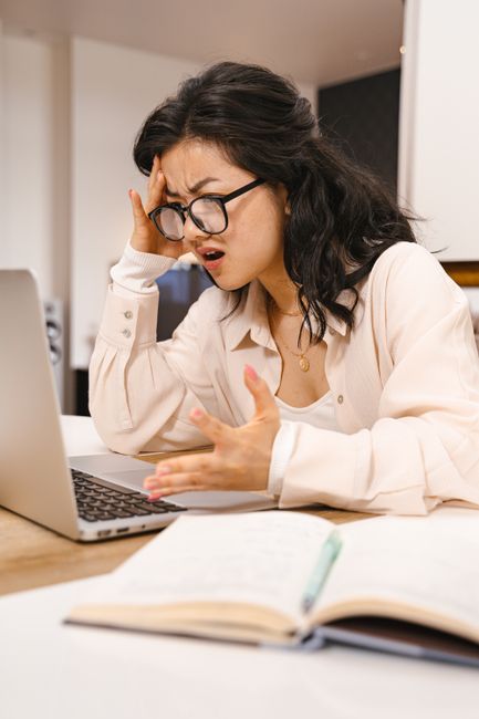 A woman with long dark hair, wearing a white shirt, sits at a desk in an office environment with a black wall background. Her facial expression shows confusion or frustration as she raises her hands w