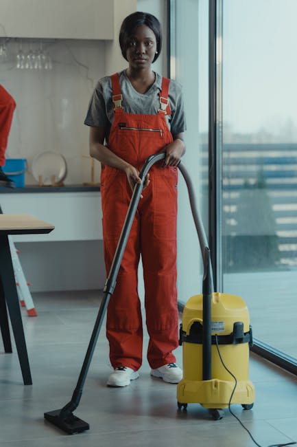 A professional cleaner, dressed in grey top and bright red overalls, stands in a modern kitchen holding a vacuum cleaner. The vacuum, with a yellow body and black accents, is positioned on a light grey tile floor, which appears clean and dust-free. The room features a sleek, contemporary design with a large glass sliding door letting in natural light, reflecting on the smooth, polished surfaces of the kitchen counters and floor. Visible in the background are minimalist white cabinetry, a glass-fronted cupboard with wine glasses, and a small wooden dining table. The overall scene emphasizes the process of surface cleaning and sanitisation as part of domestic cleaning services offered by Cleaners Kingston near Kingston's Bentall Centre.