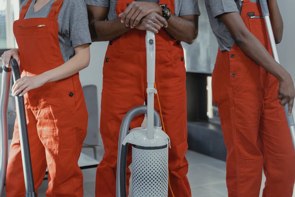 Close-up of three professional cleaning technicians standing side by side in an indoor setting, dressed in grey shirts and orange overalls. They are holding various cleaning tools including a vacuum cleaner, a mop, and a spray bottle. The vacuum cleaner, positioned centrally, features a white perforated dust container and a flexible grey hose, with the handle extending upward. The background shows a modern, well-lit room with tiled flooring and minimal furniture, indicating a deep cleaning or surface sanitisation process. Natural light illuminates the scene, highlighting the cleanliness and organized equipment used by Cleaners Kingston, a professional cleaning service near the Bentall Centre in Kingston, specializing in domestic and commercial surface cleaning.