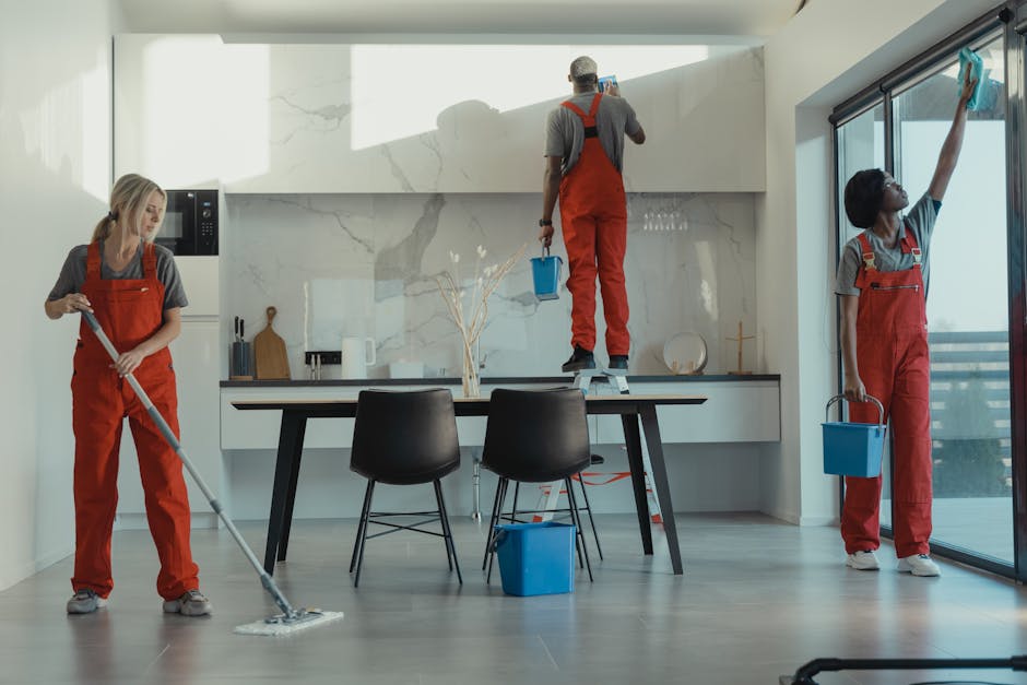 A professional cleaner, dressed in grey top and bright red overalls, stands in a modern kitchen holding a vacuum cleaner. The vacuum, with a yellow body and black accents, is positioned on a light grey tile floor, which appears clean and dust-free. The room features a sleek, contemporary design with a large glass sliding door letting in natural light, reflecting on the smooth, polished surfaces of the kitchen counters and floor. Visible in the background are minimalist white cabinetry, a glass-fronted cupboard with wine glasses, and a small wooden dining table. The overall scene emphasizes the process of surface cleaning and sanitisation as part of domestic cleaning services offered by Cleaners Kingston near Kingston's Bentall Centre.