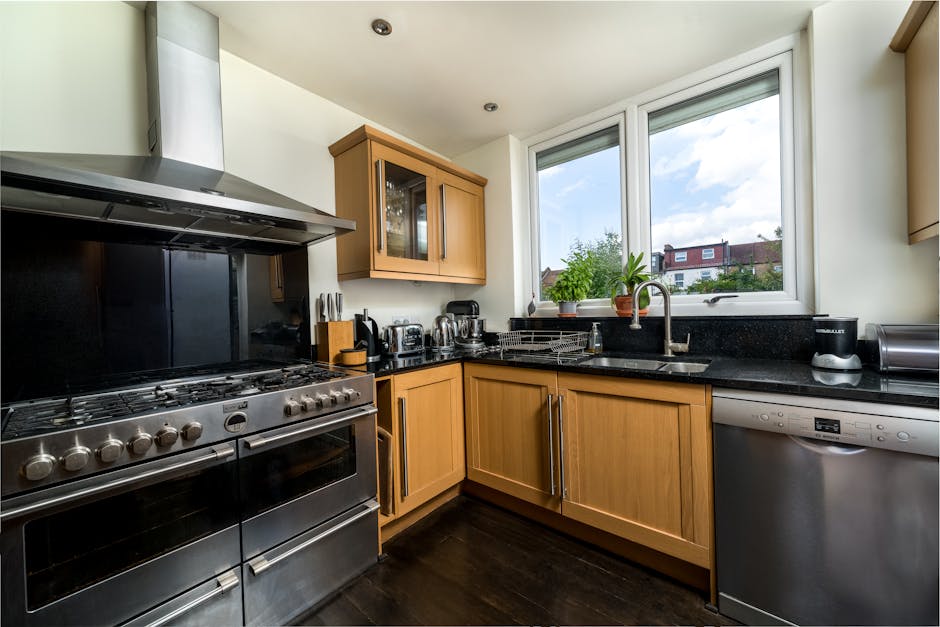 A modern kitchen featuring wooden cabinets with a natural finish, black granite countertops, and stainless steel appliances including a gas stove, oven, and dishwasher. The window above the sink allow