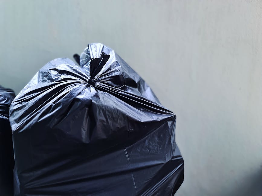 A row of outdoor wheelie bins made of plastic, positioned against a brick wall, each with a different colored lid including green, red, blue, and dark green. The bins are clean and dry, with some lids
